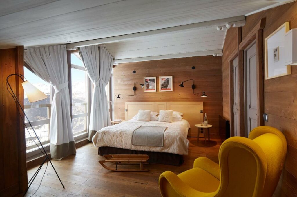 Quarto do hotel Les Trois Vallées. No meio há uma cama de casal encostada na parede, de um lado existem duas portas fechadas e, do outro lado, estão as janelas e porta para a varanda com uma vista para as montanhas cheias de neve. No canto da imagem e em frente a cama há uma poltrona amarela. O quarto é todo de madeira. Foto para ilustrar post sobre hotéis em Courchevel.