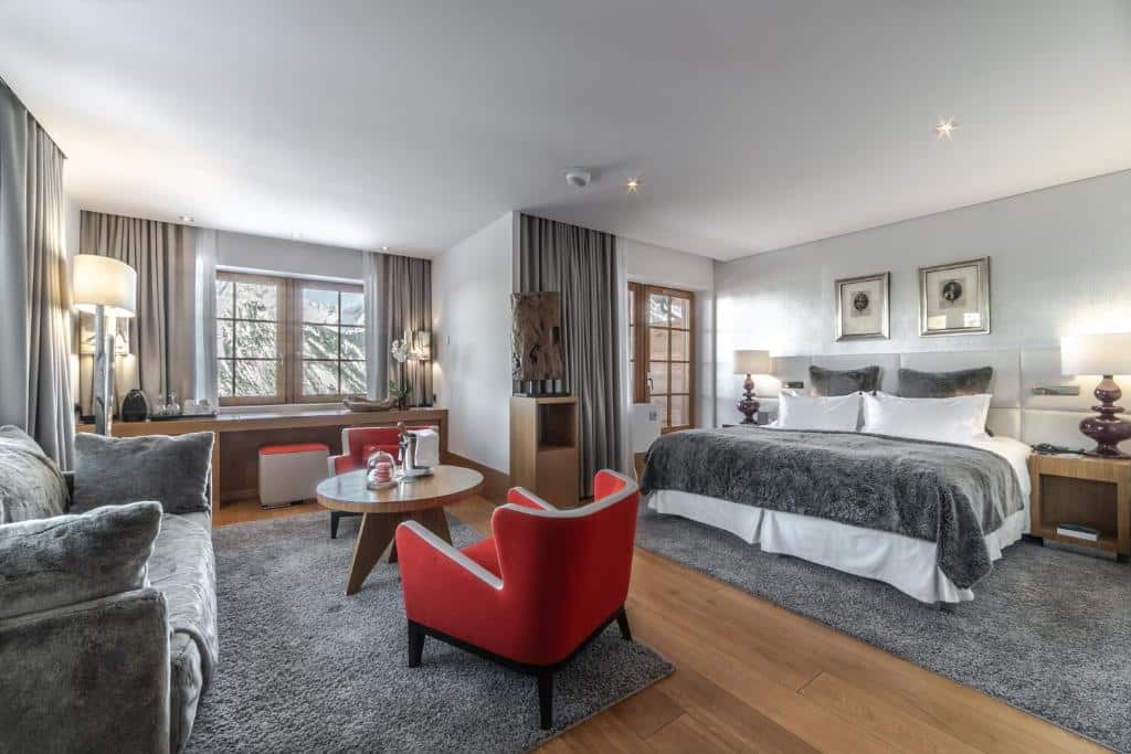 Quarto do hotel Le Strato com 63 m². A cama de casal está encostada na parede e no centro do quarto, ao lado há uma mesa com abajur e do outro lado uma porta para a varanda. Mais em frente está o sofá, duas poltronas e uma mesinha no meio deles. No canto está a janela mostrando uma montanha. Foto para ilutrar post sobre hotéis em Courchevel.