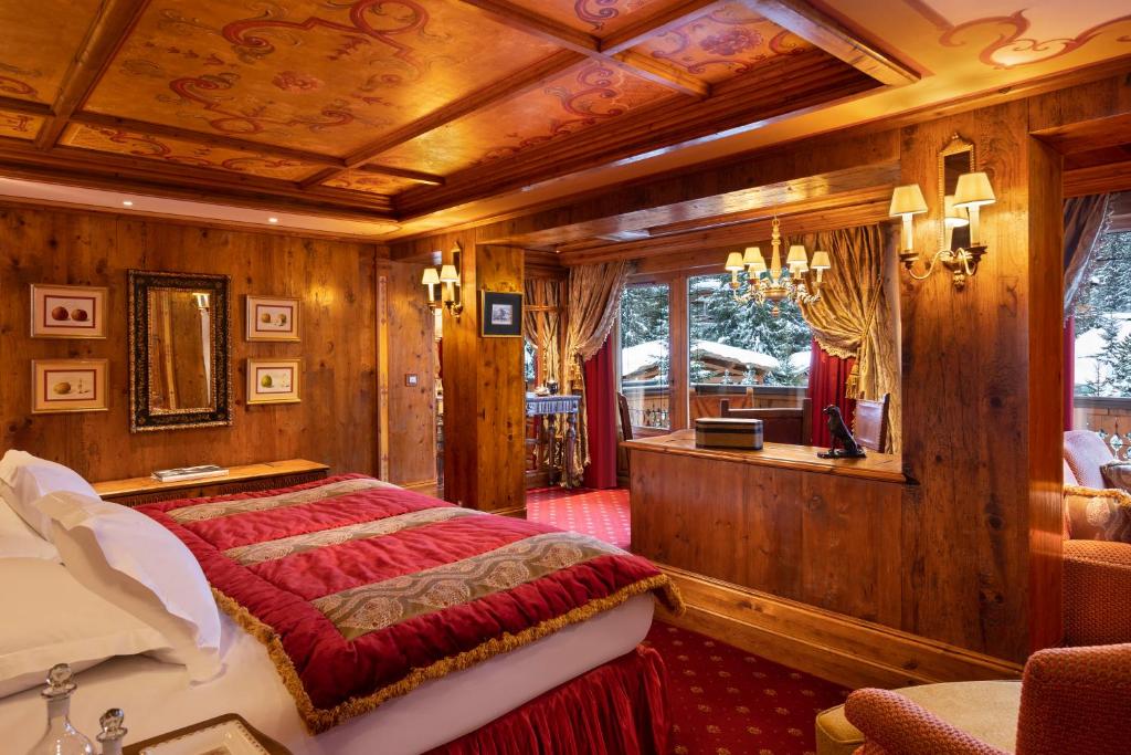 Quarto do hotel Airelles Courchevel, Les Airelles. Há uma cama de casal encostada na parede, uma poltrona ao lado e quadros e espelhos na parede. Em frente há uma divisa no quarto, separando a parte da cama do restante da acomodação. Existem janelas mostrando a vista para as árvores. O quarto é todo de madeira e possui uma decoração clássica. Foto para ilustrar post sobre hotéis em Courchevel.