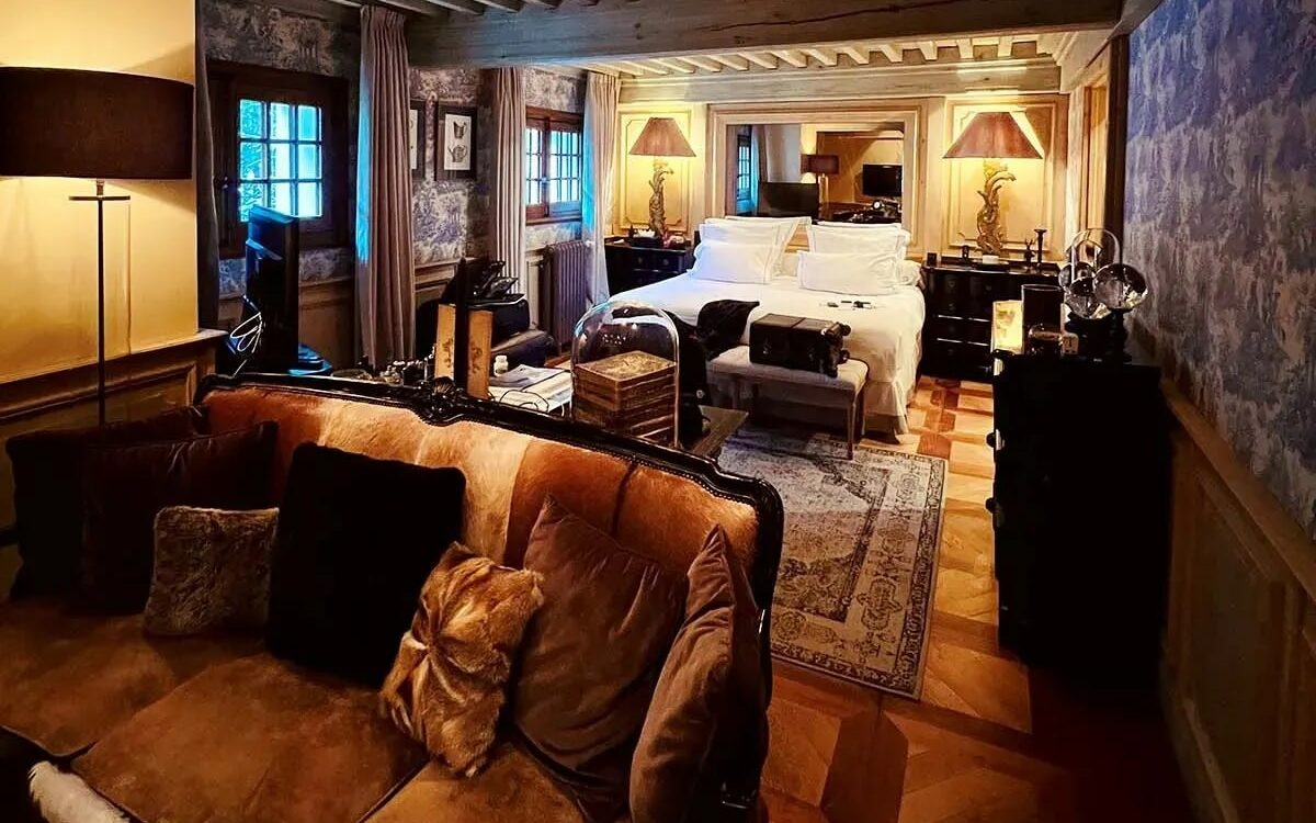 Quarto do hotel Le Saint Joseph. O quarto é cumprido e ao fundo há uma cama de casal, bem abaixo de um espelho. Há duas mesinhas com abajures aos lados da cama e um recamier na ponta. O quarto é bem decorado, possui um tapete no meio, um armário pequeno encostado na parede e iluminação amarelada. Existem janelas pequenas e cortinas. Mais para frente há um sofá. Foto para ilustrar post sobre hotéis em Courchevel.