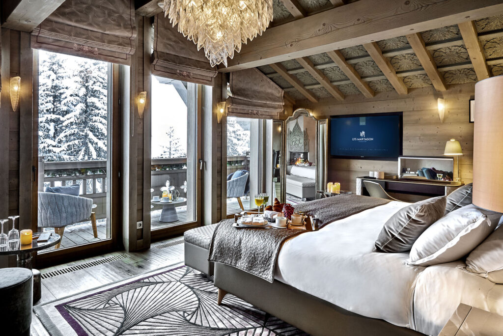 Quarto de 70m² do hotel Lys Martagon com uma cama de casal bem em frente às portas de vidro que levam até a varanda e mostram uma paisagem externa. Existe um café da manhã em cima da cama, uma televisão embutida na parede ao lado e uma mesa e cadeira no canto do quarto. Foto para ilustrar post sobre hotéis em Courchevel.