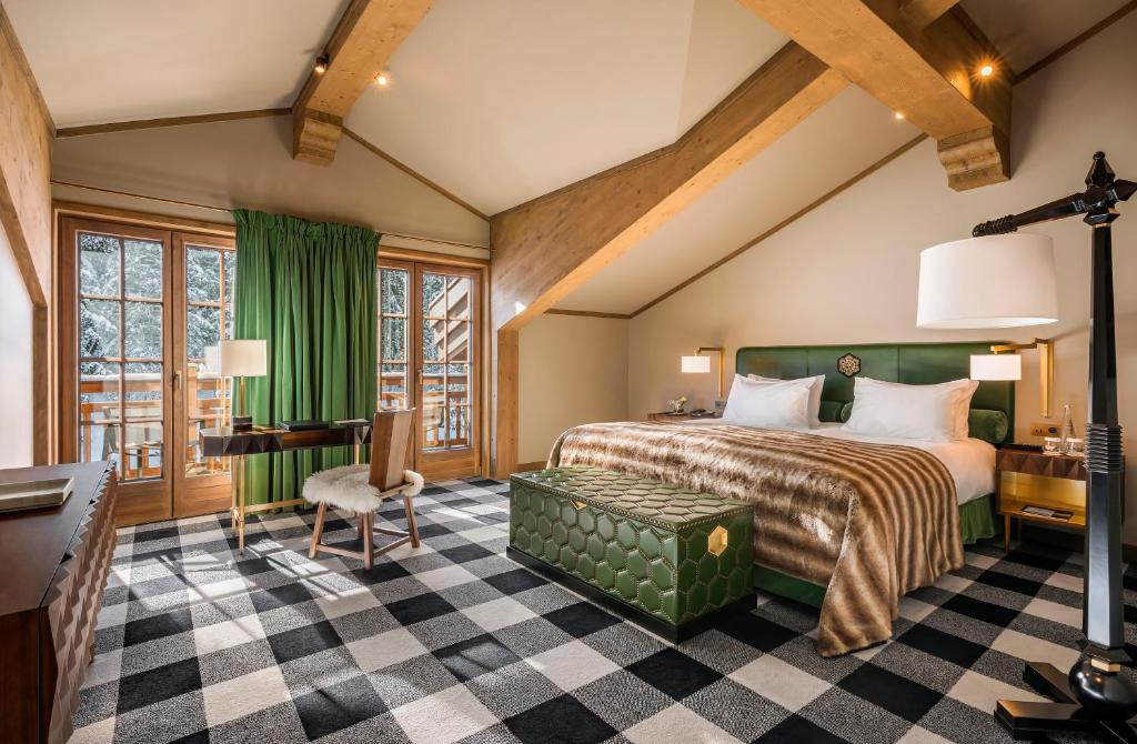 Quarto de 100 m² do hotel L'Apogée Courchevel. O chão possui um carpete todo xadrez e a cama de casal é grande e está encostada na parede, no centro do quarto. Há um baú ao pé da cama, um grande abajur no canto direito e uma mesa com cadeira do lado esquerdo. Atrás dela estão as portas que levam até a varanda. Foto ilustrar post sobre hotéis em Courchevel.