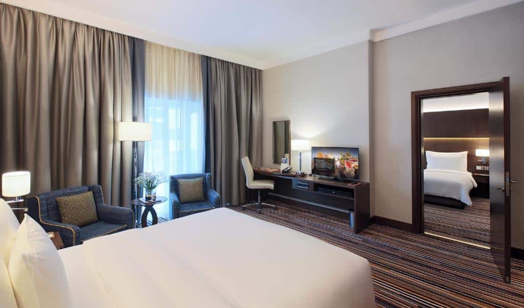 Quarto do Dusit D2 Kenz Hotel, com cama de casal, poltronas, mesa com TV e porta dando acesso a outro quarto com cama