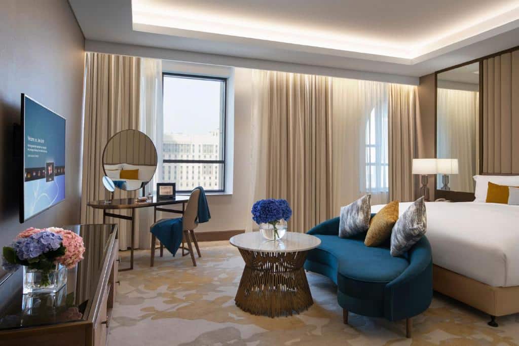 Quarto moderno do Al Jaddaf Rotana Suite Hotel com uma cama, sofá azul, escrivaninha, TV e janela grande com vista para a cidade; flores na mesa e na cômoda.