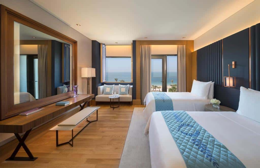 Moderno quarto do Caesars Palace Dubai com duas camas, um grande espelho, escrivaninha e vista para o mar através de grandes janelas.