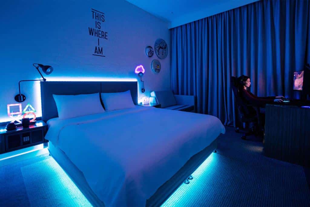 Um quarto moderno do Rove At The Park com iluminação LED azul, uma cama grande e uma pessoa usando um computador em uma mesa perto de cortinas fechadas.
