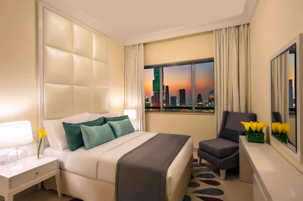 Um quarto moderno do Damac Maison Dubai Mall Street com uma cama de casal, mesas de cabeceira, uma cadeira, uma escrivaninha e uma janela que mostra o horizonte da cidade ao pôr do sol.