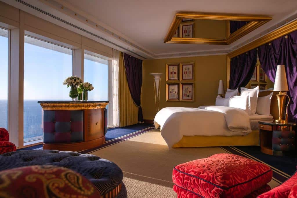 Quarto do Burj Al Arab Jumeirah com uma cama grande, espelho com moldura dourada, assentos vermelhos, janelas do chão ao teto e vista para o mar.