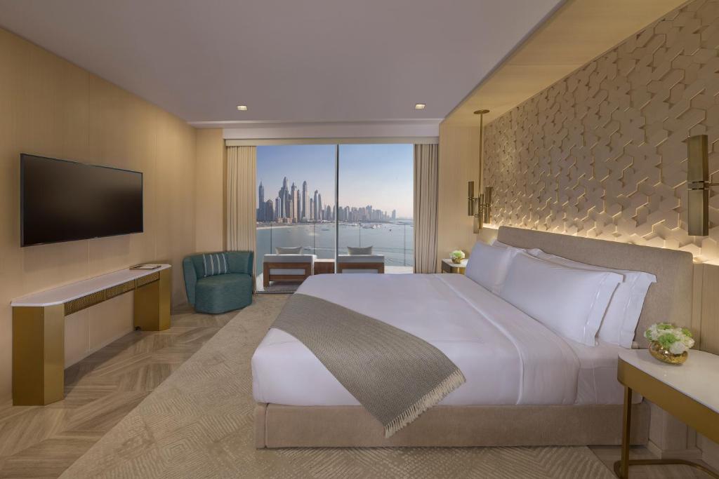 Quarto do Five Palm Jumeirah Dubai moderno com uma cama grande, TV, poltrona e vista da cidade através de janelas do chão ao teto.