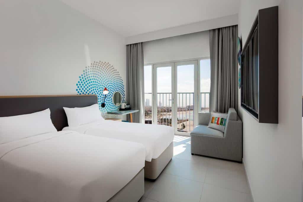 Um quarto do Rove La Mer Beach com duas camas de solteiro, um pequeno sofá perto de uma janela e uma TV na parede, com entrada de luz solar.