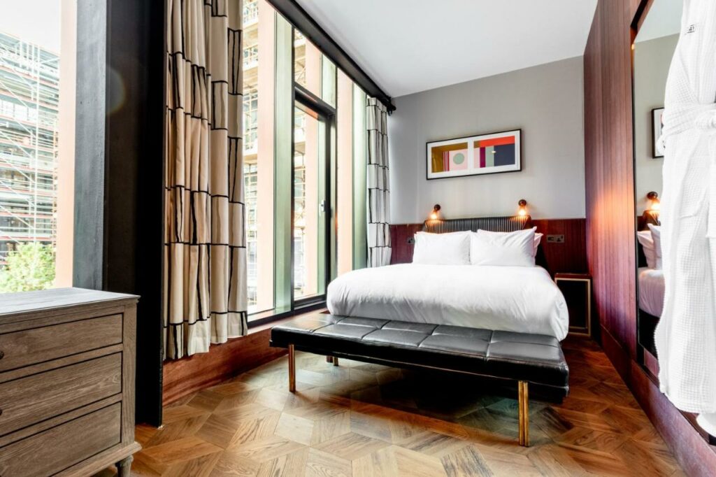 Quarto com uma janela que ocupa uma parede inteira no The Mayson  com uma cama de casal, iluminação indireta, piso de madeira e uma cômoda, para representar hotéis em Dublin.