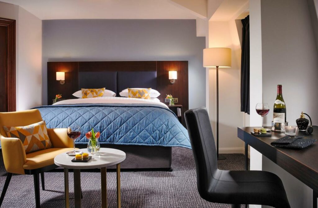 Quarto do Temple Bar Hotel com uma cama de casal, uma pequena mesa de centro com uma poltrona ao lado, uma mesa de escritório com uma cadeira, um abajur de chão e tudo decorado em tons de azul turquesa e amarelo, para representar hotéis em Dublin.