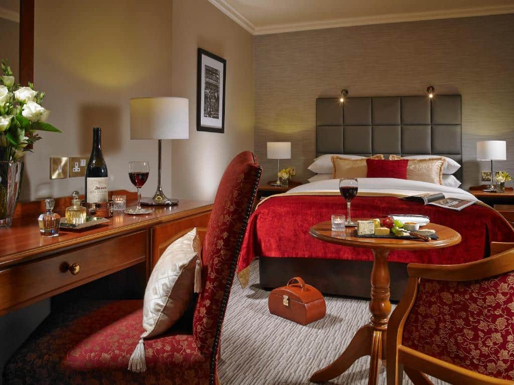 Quarto no Buswells Hotel com uma cama de casal, uma pequena mesa de centro com uma poltrona, uma mesa de escritório com uma cadeira, alguns itens de coração, duas mesinhas de cabeceira ao lado da cama com abajures, tudo decorado em tons de vermelho e dourado, para representar hotéis em Dublin.