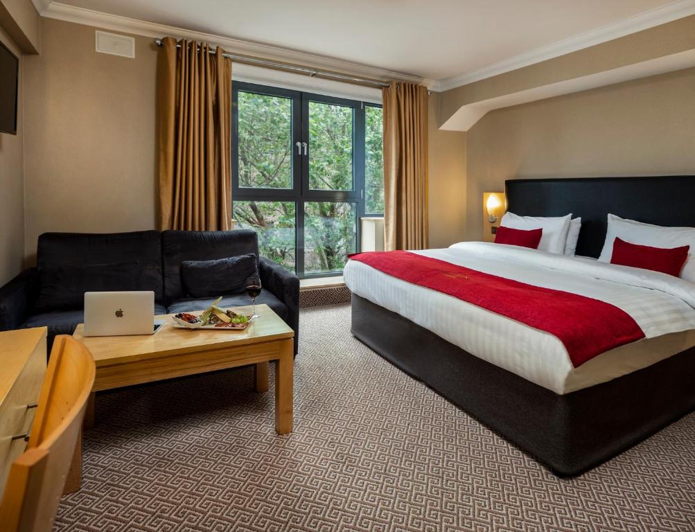 Quarto espaçoso no Academy Plaza Hotel com uma janela ampla, uma sofá, uma mesa de centro, uma cômoda e uma cama de casal, tudo em tons mais neutros, apenas a roupa de cama é vermelha, para representar hotéis em Dublin.