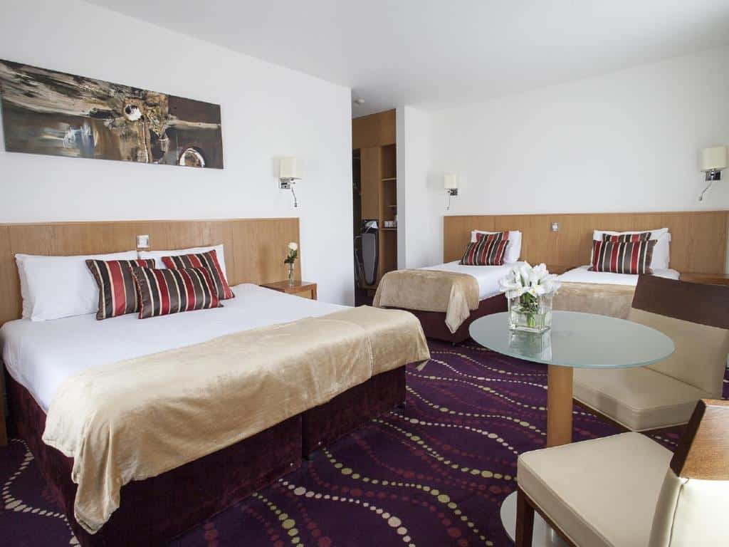 Quarto amplo no Arlington Hotel O'Connell Bridge com uma cama de casal, duas de solteiro, uma pequena mesa com duas cadeiras, tudo em tons de bege e roxo.