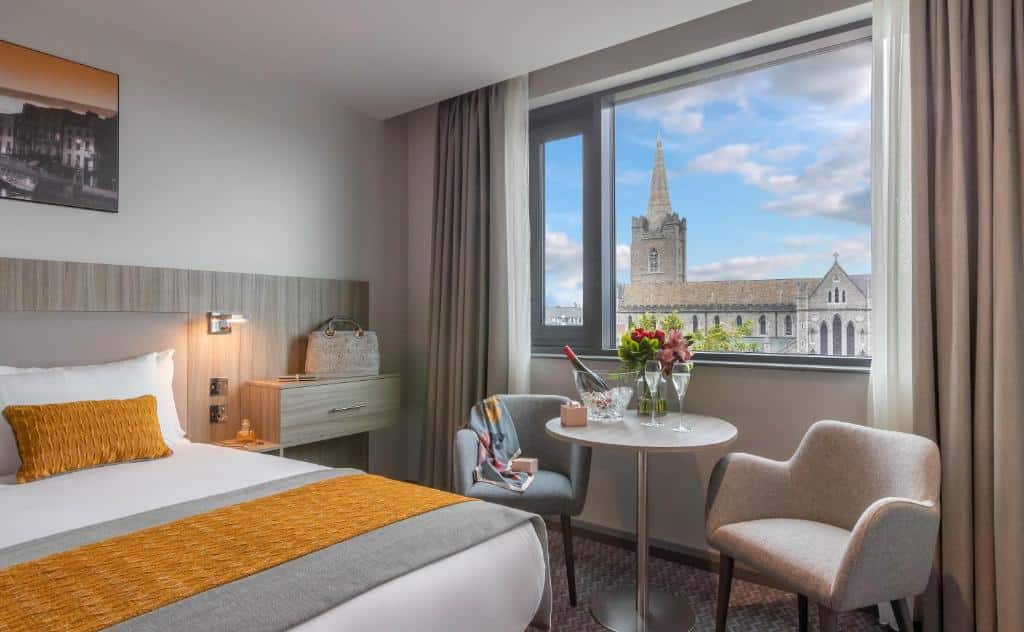 Quarto no Maldron Hotel Kevin Street com uma janela dando vista para a cidade, uma mesinha central com duas poltronas, uma cama de casal e uma cabeceira com gavetas e iluminação indireta.