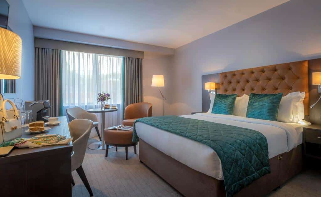Quarto do Grand Canal Hotel com uma cama de casal, uma janela, duas poltronas e uma pequena mesinha, uma mesa de escritório com uma cadeira, um espelho e dois abajures de chão, para representar os hotéis em Dublin.