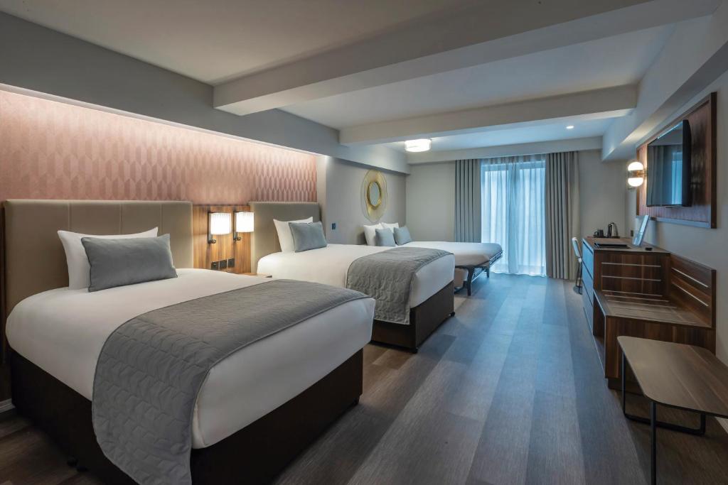 Quarto no Riu Plaza The Gresham Dublin com três camas, piso de madeira, escrivaninha, TV de tela plana e uma janela com cortinas que permitem a entrada de luz natural, para representar hotéis em Dublin.