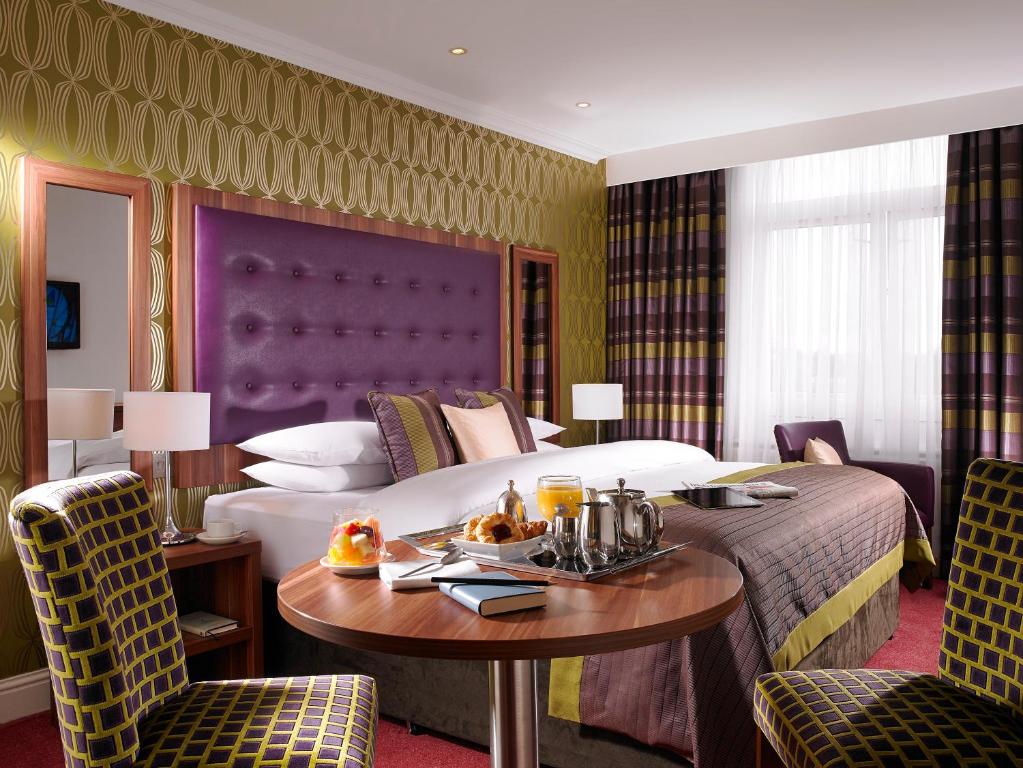 Quarto do Dublin Skylon Hotel com uma cama de casal, com uma cabeceira roxa e almofadas sob a cama, uma janela com cortinas e uma mesa redonda de madeira com duas cadeiras estofadas, para representar hotéis em Dublin.