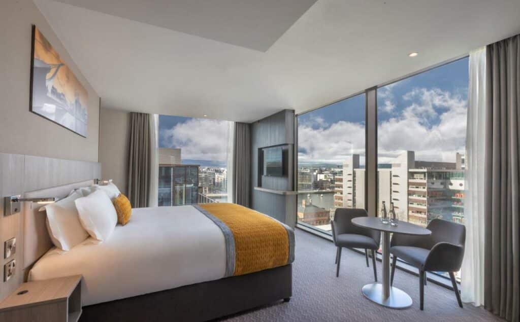 Quarto com vista para a cidade no The Samuel Hotel com uma cama de casal, uma televisão, uma pequena mesa com dois lugares, tudo decorado em tons de amarelo, cinza e branco.