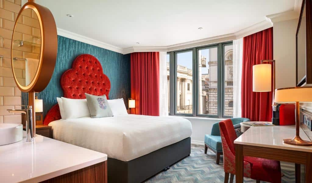 Quarto no Hard Rock Hotel Dublin com uma janela ampla com cortinas vermelhas, uma poltrona, uma mesa de trabalho com uma cadeira, uma televisão, uma cama de casal, tudo decorado em tons de azul turquesa, vermelho e branco.