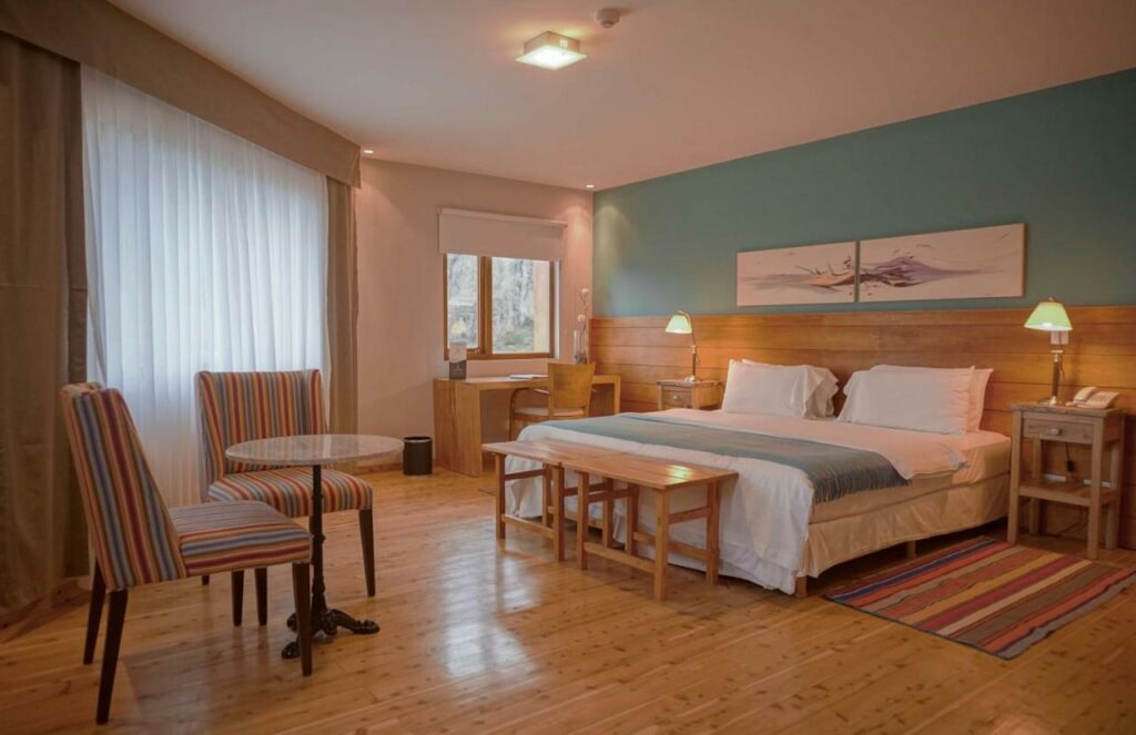 Quarto do Destino Sur Hotel & Spa de Montaña com cama de casal do lado direito da imagem com uma cômoda em cada lado com luminária do lado direito da cama uma mesa de trabalho com cadeira. No pé da cama um banco de madeira e em frente duas cadeiras estofadas com mesinha, para representar hotéis em El Chalten.