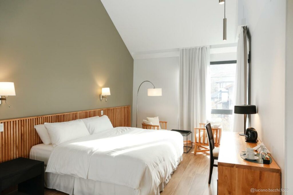 Quarto do Kaulem Hotel Boutique com cama de casal do lado esquerdo da imagem em frente a cama uma cômoda com cadeira, para representar hotéis em El Chalten.