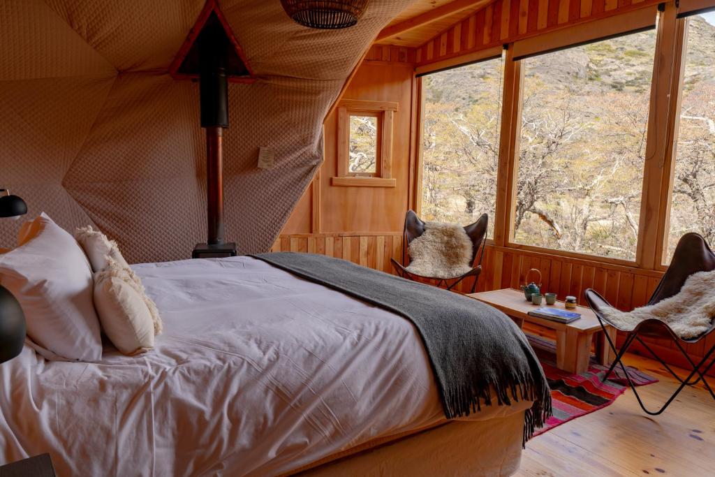 Quarto aconchegante no Patagonia Eco Domes com janelas grandes, uma cama de casal, duas cadeiras com mantas de pele e vista para as árvores do lado de fora, para representar hotéis em El Chalten.