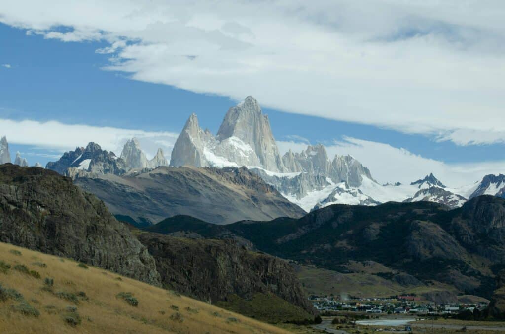 Montanhas com picos cobertos de neve se erguem atrás de uma pequena cidade e colinas gramadas sob um céu parcialmente nublado, em El Chalten.