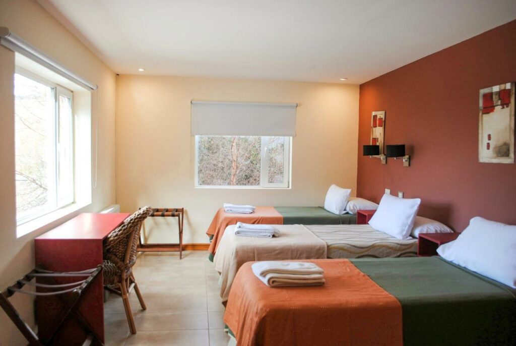 Quarto familiar do Hotel Lunajuim com três camas de solteiro do lado direto da imagem em frente as camas uma mesa de trabalho com cadeira, para representar hotéis em El Chalten.
