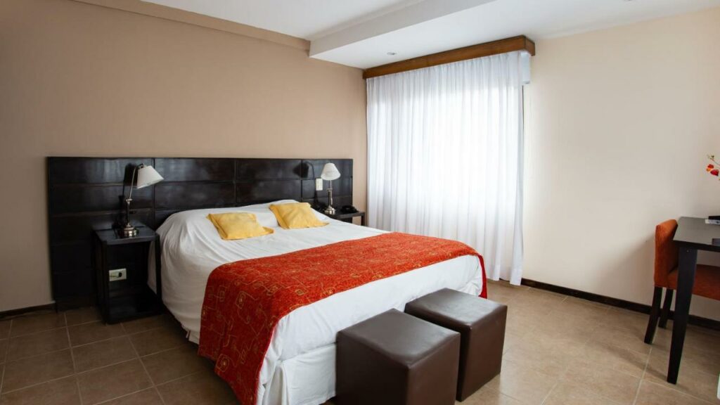 Quarto do Hotel Poincenot  com cama de casal do lado esquerdo da imagem no centro em cada lado da cama uma cômoda com luminária e no pé da cama dois puffs, para representar hotéis em El Chalten.