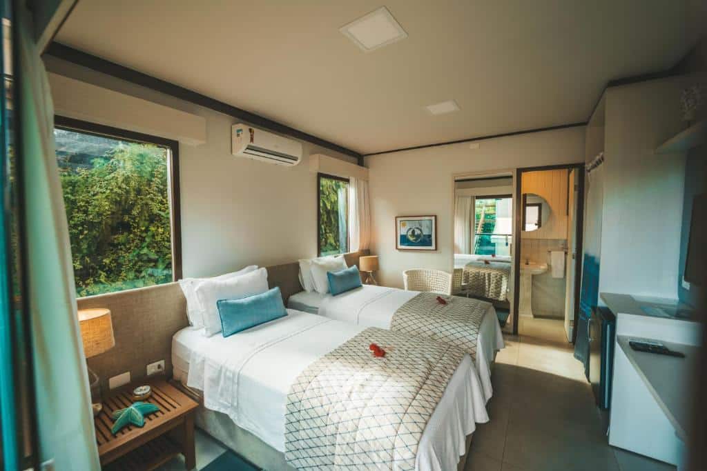 Quarto do Tesouro de Noronha. Duas camas de solteiro nomeio, em cima um ar-condiconado, de cada lado uma mesinha com um abajur, e duas janelas grandes. De frente, uma mesa com um frigobar e uma televisão. No fundo a porta do banheiro e um espelho. Foto para ilustrar post sobre hotéis em Fernando de Noronha.