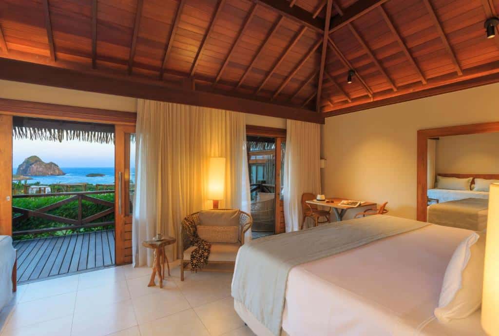 Quarto do NANNAI Noronha Solar Dos Ventos. Uma cama de casal no lado direito, de frente uma poltrona e uma mesa com duas cadeiras. Atrás duas portas para a sacada do quarto e vista para o mar. Foto para ilustrar post sobre hotéis em Fernando de Noronha.