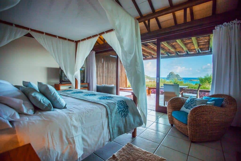 Quarto da Pousada Maravilha. Uma cama de casal do lado esquerdo, do lado direito uma poltrona, no fundo do lado esquerdo uma televisão. De frente pra cama a porta de vidro para a sacada, com hidro, mesa, cadeiras e vista para o mar. Foto para ilustrar post sobre hotéis em Fernando de Noronha.