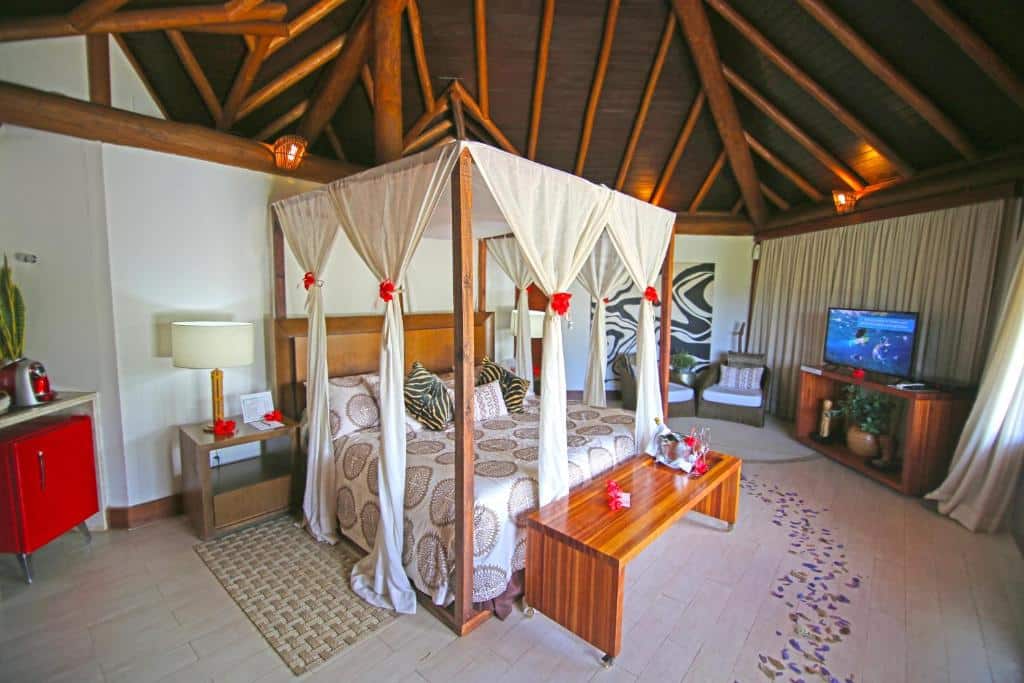 Quarto da Pousada Triboju. Uma cama de casal no meio, com um banco na frente. No lado direito, duas poltronas uma raque e uma televisão. Do lado esquerdo um frigobar e uma cafeteira e uma cômoda com abajur do lado da cama. Foto para ilustrar post sobre hotéis em Fernando de Noronha.