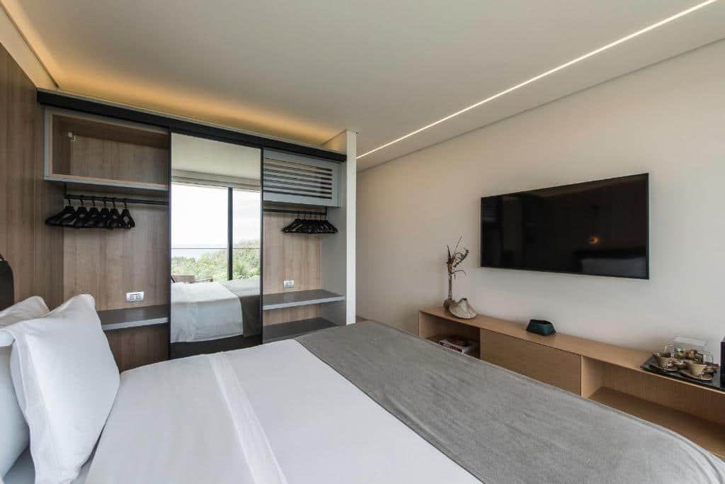 Quarto da Pousada Maria Flor. Uma cama de csal do lado esquerdo, de frente uma raque com uma televisão. Atrás, no fundo, um guarda-roupa com espelho e cabides. Foto para ilustrar post sobre hotéis em Fernando de Noronha.