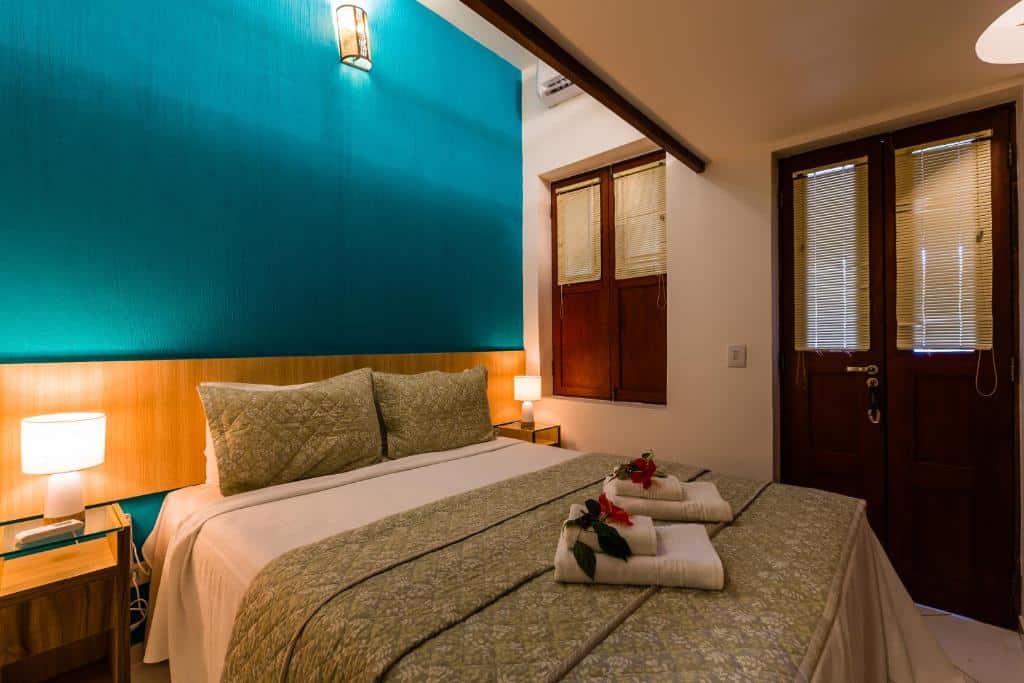 Quarto da Casa Conceição. Uma cama de casal no meio, de cada lado uma cômoda com um abajur. No lado direito a porta e a janela do quarto. Foto para ilustrar post sobre hotéis em Fernando de Noronha.