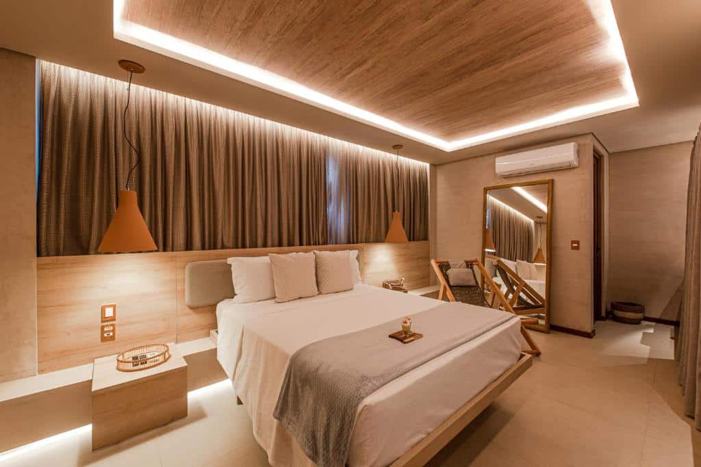 Quarto da POUSADA DO MIRANTE. Uma cama de casal no meio, do lado direito da cama uma cadeira de madeira, um espelho e o ar-condicionado em cima. De cada lado da cama, pendurado um lustre, e atrás a cortina do quarto. Foto para ilustrar post sobre hotéis em Fernando de Noronha.