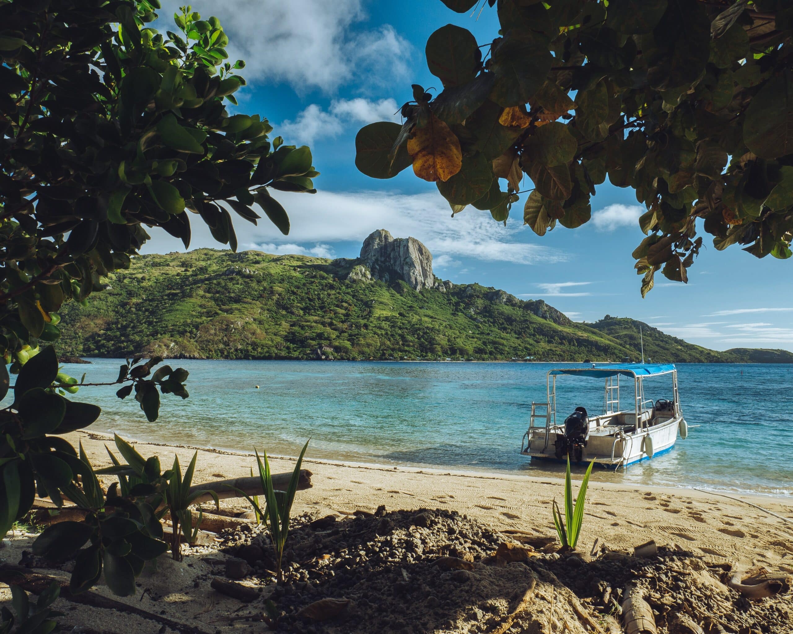Vista da praia em Kuata, Fiji com areia a frente e barco parado na beira do mar com mar azul ao fundo montanhas durante o dia.
