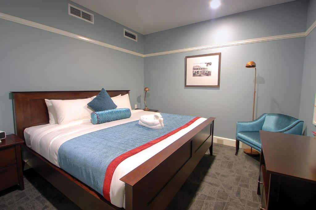 Quarto do Grand Pacific Hotel com cama de casal com duas cômodas de madeira ao lado com cobertas brancas e azuis, poltrona azul em frente a cama com uma cômoda de madeira.