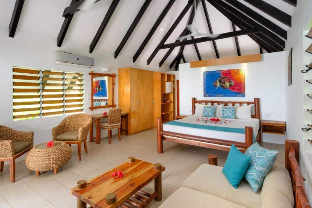 Quarto do Tropica Island Resort-Adults Only com cama de casal, duas cômodas de madeira ao lado da cama, guarda-roupa do lado esquerdo e ao lado uma mesa de trabalho com cadeira em frente a cama uma pequena sala de estar com sofá bege, mesa de centro e duas poltronas de palhas.
