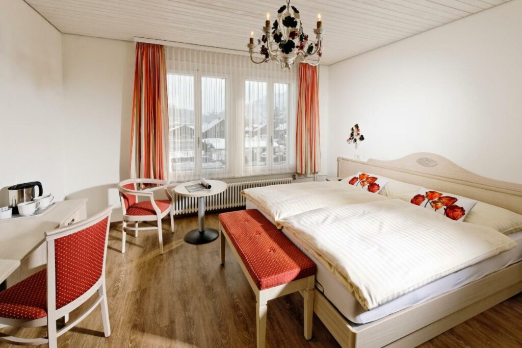 Quarto do Hotel Beausite com cama de casal do lado direito imagem no pé da cama um banco estofado e em frente a cama uma mesa de trabalho com cadeira do lado esquerdo da cama uma mesa redonda com cadeira. Representa hotéis em Interlaken.