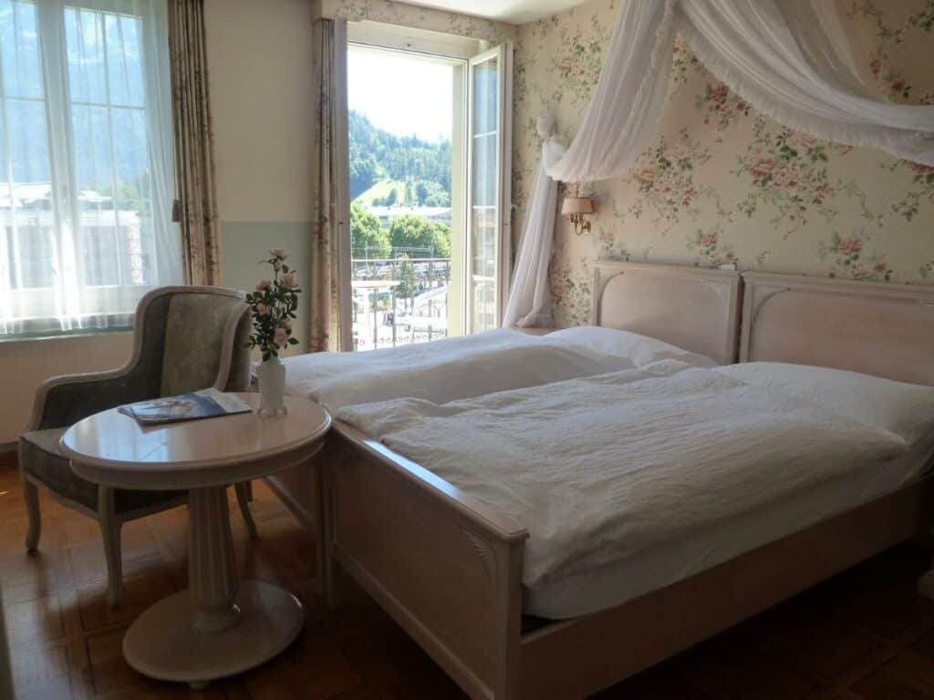 Quarto do Hotel Central Continental com cama de casal do lado direito da imagem no centro do quarto nos pés da cama uma mesa redonda com uma cadeira e do lado direito da cama um porta que dá acesso a varanda.