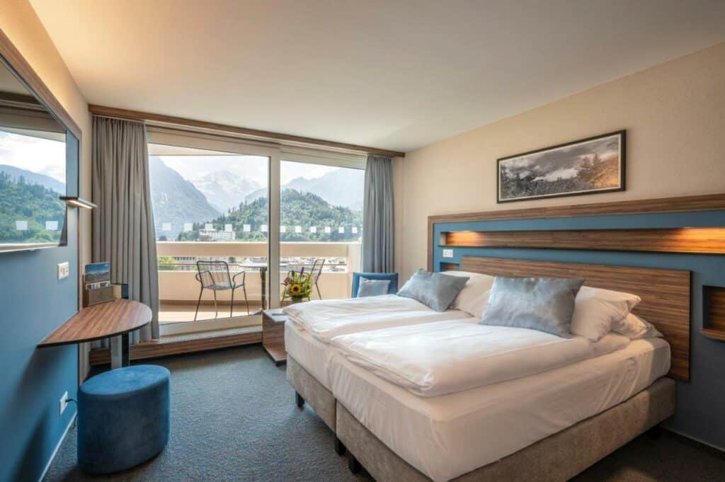 Quarto do Metropole Swiss Quality Hotel com cama de casal do lado direito da imagem no centro do quarto em frente a cama a TV presa na parede do lado direito da cama uma poltrona com uma mesa e ao fundo uma porta de vidro que dá acesso a varanda. Representa hotéis em Interlaken.