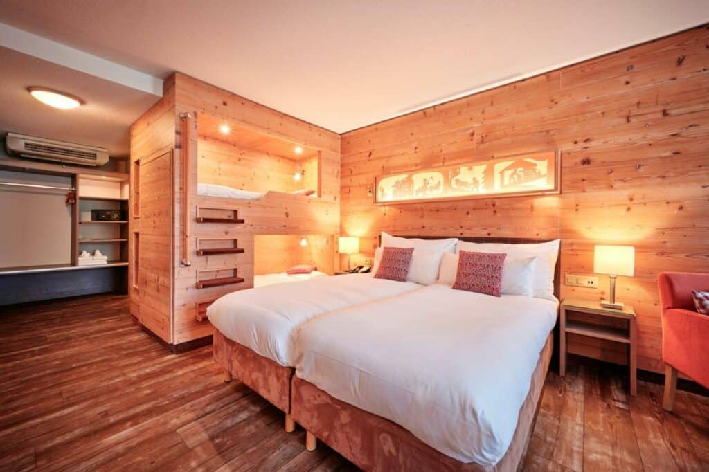 Imagem do quarto do Hotel Interlaken com cama de casal do lado no centro do ambiente em cada lado do quarto uma cômoda com luminária e do lado direito da cama ao lado da cômoda uma beliche. Representa hotéis em Interlaken.
