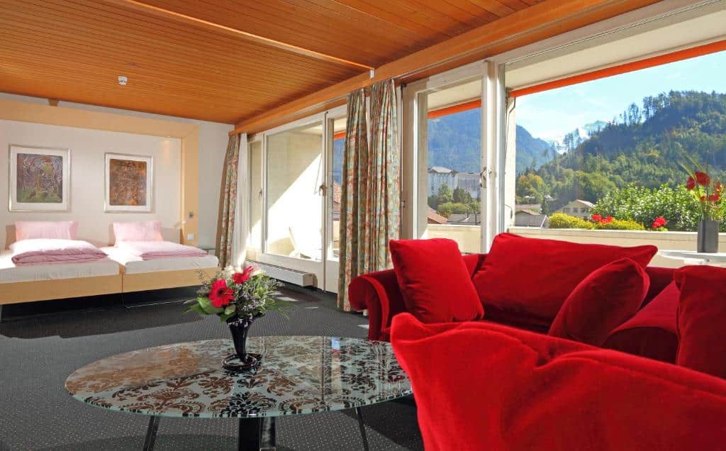 Quarto do Stella Swiss Quality Hotel  com um sofá do lado direto da imagem e uma mesa redonda e ao fundo uma cama de casal do lado esquerdo da cama uma porta de vidro que dá acesso a varanda. Representa hotéis em Interlaken.
