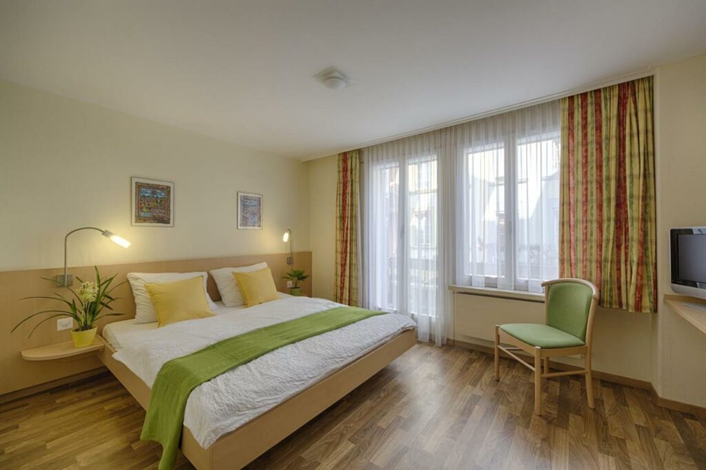 Imagem do quarto do Hotel Blume Interlaken com cama de casal do lado esquerdo da imagem do lado esquerdo da cama a frente uma cadeira e atrás da cadeira uma TV em cima de uma cômoda. Representa hotéis em Interlaken.