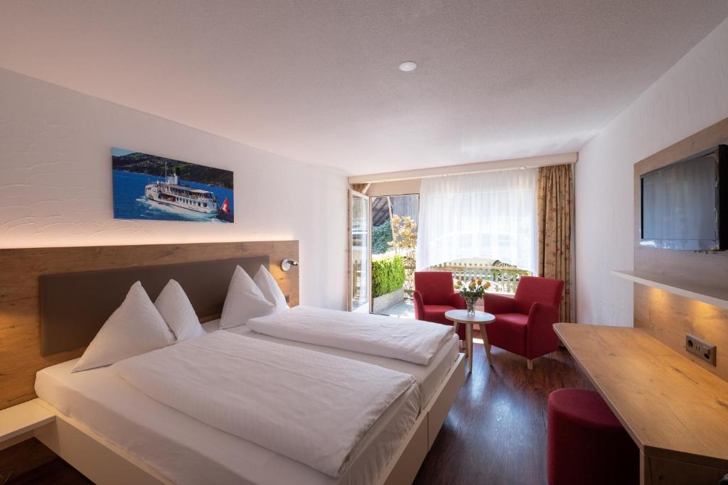 Quarto do Hotel Chalet Swiss com cama de casal do lado esquerdo da imagem em frente a cama uma cômoda com a TV presa na parede do lado esquerdo do ambiente duas poltronas com uma mesa no centro e ao fundo uma porta que dá acesso a varanda. Representa hotéis em Interlaken.