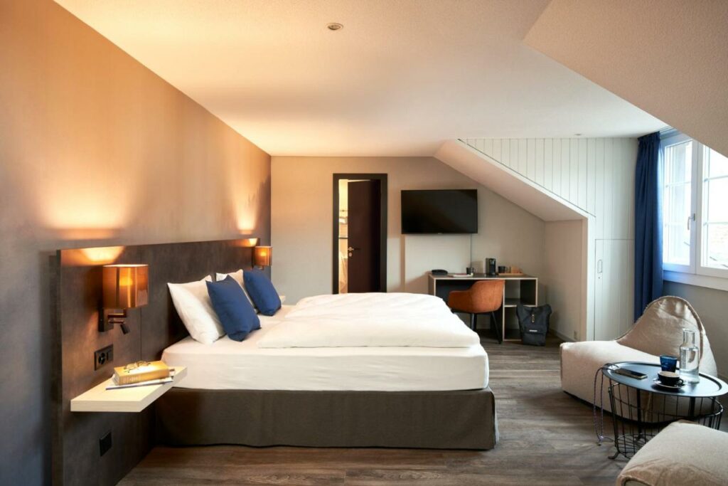 Imagem do quarto do The Hey Hotel  com cama de casal do lado esquerdo da imagem em frente a cama um puff e do lado esquerdo do puff uma mesa redonda do lado esquerdo da cama em frente ao lado uma mesa de trabalho com cadeira e em cima a TV presa. Representa hotéis em Interlaken.