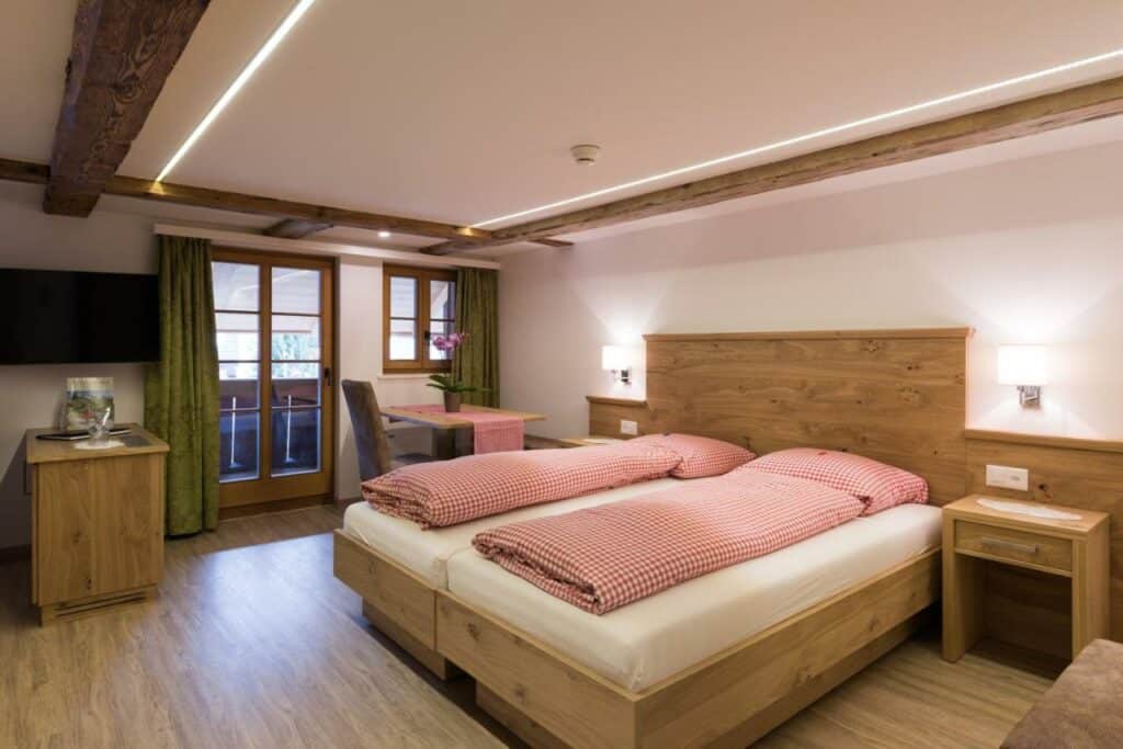 Quarto do Hotel Restaurant Hirschen com cama de casal do lado direito da imagem no centro do quarto em cada lado da cama uma cômoda do lado direito da cama uma mesa com cadeira e ao fundo uma cômoda e uma TV presa na parede. Representa hotéis em Interlaken.